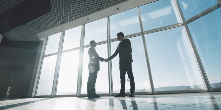 Two young businessmen are shaking hands with each other standing against panoramic windows.