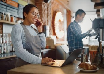 Latin American Coffee Shop Employee Accepts a Pre-Order on a Mobile Phone Call and Writes it Down on Laptop Computer in a Cozy Cafe. Restaurant Manager Browsing Internet and Talking on Smartphone.