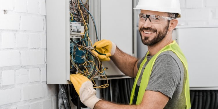 smiling handsome electrician repairing electrical box with pliers in corridor and looking at camera